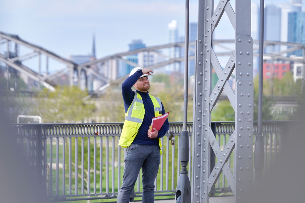 Alexander Michel mit Schutzhelm und Warnweste auf einer Stahlfachwerkbrücke, im Hintergrund die Frankfurter Skyline
